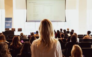 A woman in an audience standing to see what is projected on a screen.