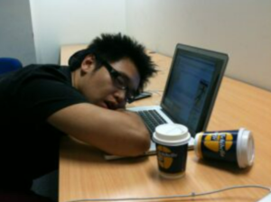 A person asleep on their laptop keyboard with an empty cup of coffee knocked over.