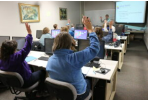 Audience seated at computer workstations, with some hands raised.