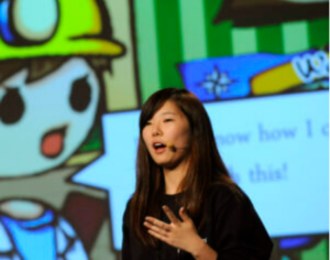A speaker addressing a group, with a cartoon figure in the background.