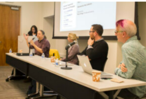 F our people at front of a room in a panel presentation, with a moderator at the side.