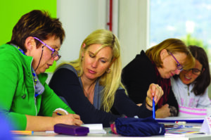 Four women working at a table, two groups of two, with each group focused on their documents.