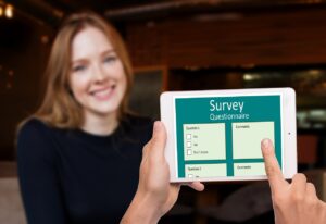 A person holding ipad with Survey on the screen; woman in background smiling.